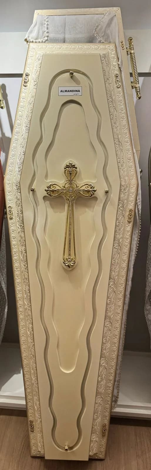 Urna funerária Almandina Delicata em acabamento claro com relevo ornamental e crucifixo dourado.
