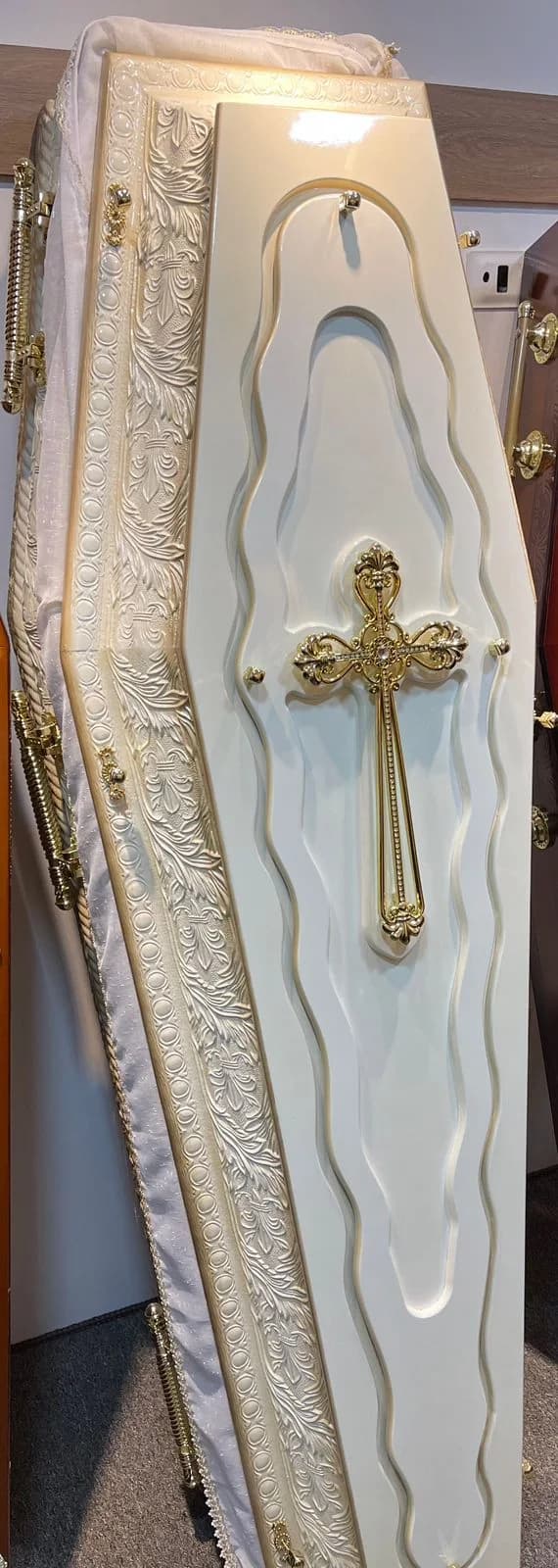 Urna funerária em acabamento marfim com relevo ornamental e crucifixo central.